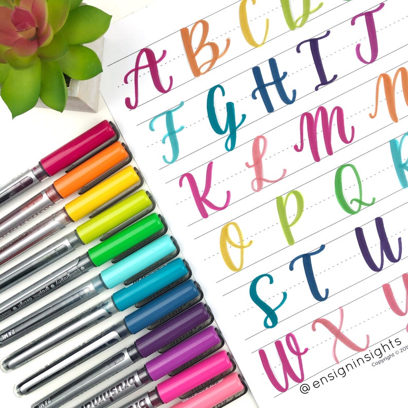 Peut inclure: Une feuille de papier avec l'alphabet &eacute;crit en cursive en diff&eacute;rentes couleurs. Les lettres sont &eacute;crites sur du papier lign&eacute;. Un ensemble de stylos color&eacute;s se trouve sur le c&ocirc;t&eacute; gauche de l'image.