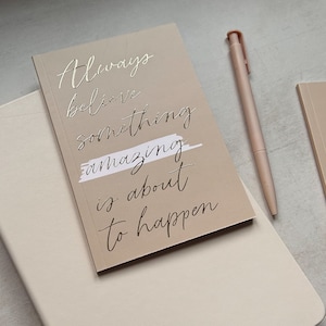 May include: A beige notebook with the text "Always believe something amazing is about to happen" written in silver script. The notebook is sitting on a beige surface with a pink pen.