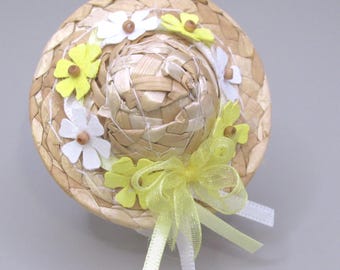 May include: A miniature straw hat with a woven design, adorned with white and yellow daisy-shaped flowers. A yellow ribbon bow with white ribbon accents adds a decorative touch. The hat is a charming accessory.