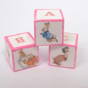 May include: Four white wooden alphabet blocks with pink edges. Each block features a letter and a colourful illustration of a bunny or duck. The blocks are stacked, creating a playful arrangement.
