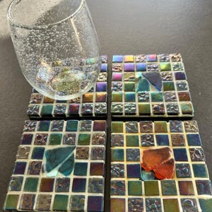 May include: Four square mosaic tile coasters with iridescent glass tiles in shades of green, blue, purple, and gold. A clear glass tumbler with bubbles rests on one coaster. Each coaster has a unique colored glass accent.