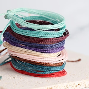 May include: A collection of colourful waxed cord bracelets in shades of teal, burgundy, purple, and pink. Each bracelet is knotted and ready to wear. The bracelets are stacked on a neutral-coloured, textured surface.