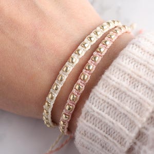 Peut inclure: Deux bracelets avec des perles dorées sur une ficelle blanche et rose. Les bracelets sont portés au poignet.