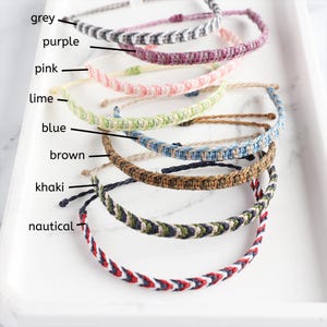 Könnte beinhalten: Eine Auswahl handgefertigter gewebter Armbänder in verschiedenen Farben, darunter Grau, Lila, Rosa, Limette, Blau, Braun, Khaki und Marine. Jedes Armband wird auf einer weißen Oberfläche präsentiert, um die verschiedenen Farboptionen hervorzuheben.