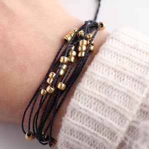 May include: A black string bracelet with multiple strands and gold beads. The bracelet is worn on a person's wrist.