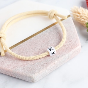 May include: A cream-coloured rope bracelet with a white cube bead featuring the letter "K" in black. The bracelet is knotted at the ends and rests on a pink and white surface with a gold accent.