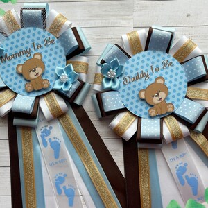 May include: Two blue and white baby shower sashes with brown and gold ribbon. One sash says "Mommy to Be" and the other says "Daddy to Be". Both sashes feature a brown teddy bear and blue footprints with the text "It's a Boy!"