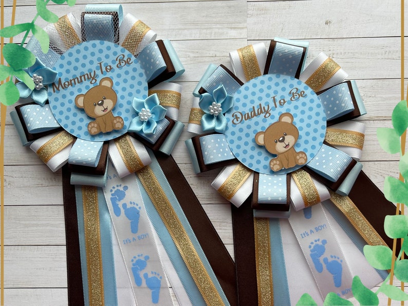 May include: Two blue and white baby shower sashes with brown and gold ribbon. One sash says "Mommy to Be" and the other says "Daddy to Be". Both sashes feature a brown teddy bear and blue footprints with the text "It's a Boy!"