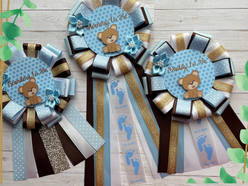 May include: Three blue and white baby shower sashes with brown and gold ribbon. The sashes have a blue polka dot background with a brown teddy bear and the text "Mommy To Be", "Daddy To Be", and "Grandma To Be".
