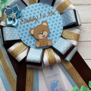 May include: A blue and white polka dot ribbon with a brown ribbon and gold glitter ribbon. The center of the ribbon has a blue circle with white polka dots and the text "Daddy To Be". A brown teddy bear with a white face is in the center of the circle.