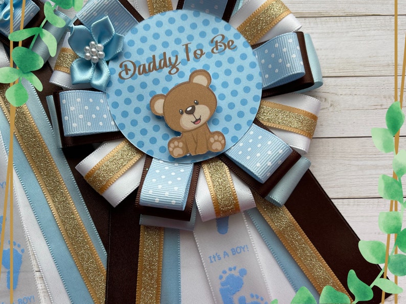 May include: A blue and white polka dot ribbon with a brown ribbon and gold glitter ribbon. The center of the ribbon has a blue circle with white polka dots and the text "Daddy To Be". A brown teddy bear with a white face is in the center of the circle.