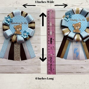 May include: Two blue and brown baby shower sashes with a teddy bear design. The sashes are 4 inches wide and 6 inches long. One sash says "Grandma To Be" and the other says "Daddy To Be".