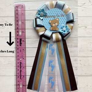 May include: Blue and brown mommy-to-be sash with a blue and white polka dot circle that says "Mommy To Be" and a brown teddy bear. The sash is 9 inches long and has blue footprints and the text "It's a Boy" printed on it.