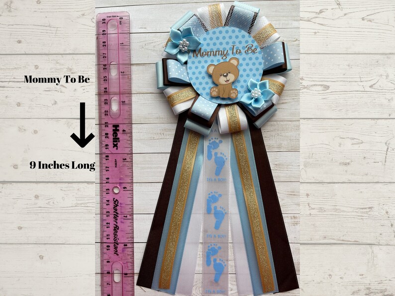 May include: Blue and brown mommy-to-be sash with a blue and white polka dot circle that says "Mommy To Be" and a brown teddy bear. The sash is 9 inches long and has blue footprints and the text "It's a Boy" printed on it.