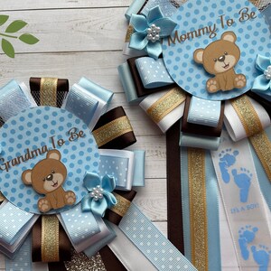 May include: Two blue and brown baby shower sashes with a teddy bear design. The sashes say "Mommy to Be" and "Grandma to Be".