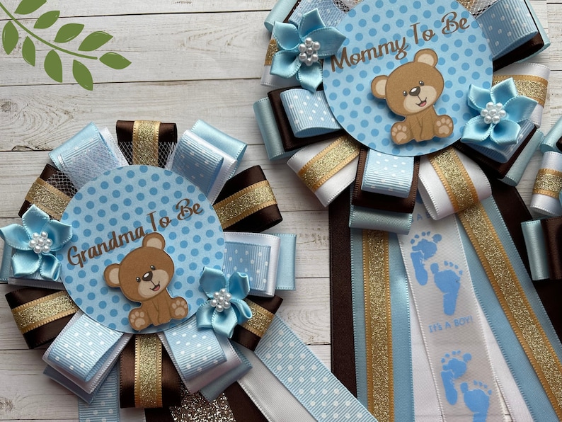 May include: Two blue and brown baby shower sashes with a teddy bear design. The sashes say "Mommy to Be" and "Grandma to Be".