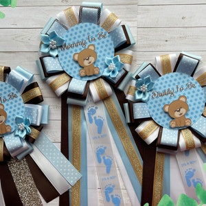May include: Three blue and white baby shower sashes with brown and gold ribbon. The sashes say "Mommy to Be", "Daddy to Be", and "Grandma to Be". Each sash has a blue and white polka dot circle with a brown teddy bear.