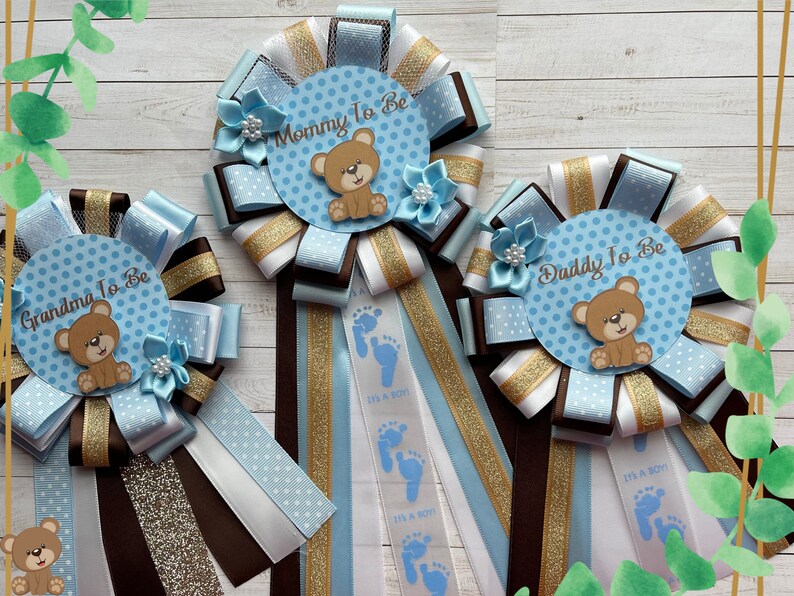 May include: Three blue and white baby shower sashes with brown and gold ribbon. The sashes say "Mommy to Be", "Daddy to Be", and "Grandma to Be". Each sash has a blue and white polka dot circle with a brown teddy bear.