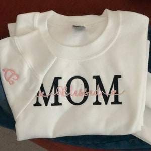Puede incluir: Sudadera blanca con texto bordado en negro que dice "MOM" con la palabra "blessed" en cursiva rosa entre la "O" y la "M".