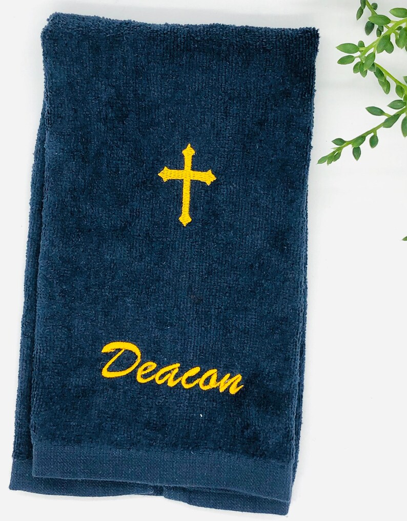 Puede incluir: Una toalla de mano azul oscuro con una cruz bordada en oro y el nombre "Deacon" en oro.