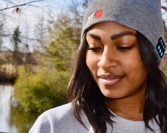 May include: A woman wearing a gray beanie with the word "LOVE" and a red heart embroidered on the front. The beanie has a built-in Bluetooth speaker with blue lights.