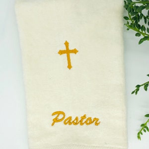 Puede incluir: Toalla de mano blanca con una cruz bordada en oro y la palabra "Pastor" en cursiva.