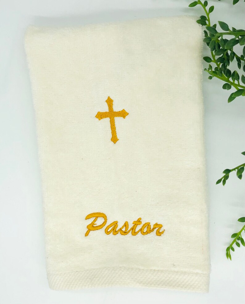 Puede incluir: Toalla de mano blanca con una cruz bordada en oro y la palabra "Pastor" en cursiva.