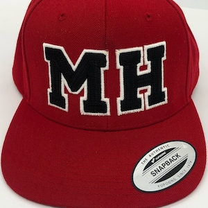 May include: Red baseball cap with a black and white embroidered "MH" logo. The cap has a curved brim and a snap closure. The back of the cap has a silver label that reads "The Authentic Yupoong Snapback Yupoong Since 1974."