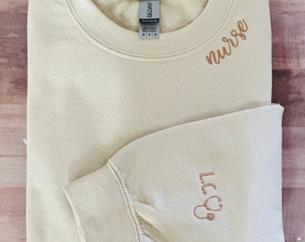 May include: A light beige sweatshirt with the word "nurse" embroidered on the front and a stethoscope with the word "L.C.N." embroidered on the sleeve.