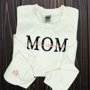 Puede incluir: Sudadera blanca con texto bordado en negro que dice "MOM" con la palabra "Blessed" en cursiva rosa debajo.