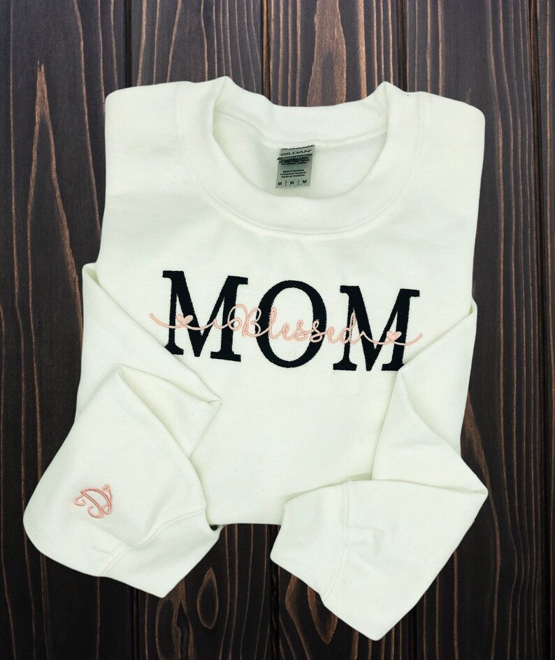 Puede incluir: Sudadera blanca con texto bordado en negro que dice "MOM" con la palabra "Blessed" en cursiva rosa debajo.
