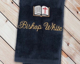 May include: A dark blue towel with gold embroidery that reads "Bishop White" and a small embroidered image of an open book with a red cross.