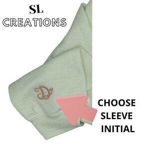 Puede incluir: Una sudadera blanca con una letra "D" bordada en rosa en la manga. El texto "CHOOSE SLEEVE INITIAL" es visible en el lado derecho de la imagen.