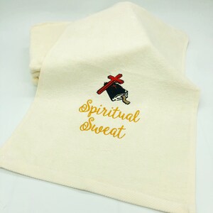 May include: A white hand towel with gold embroidery that reads "Spiritual Sweat". A small black and red illustration of a book with a cross on top is above the text.