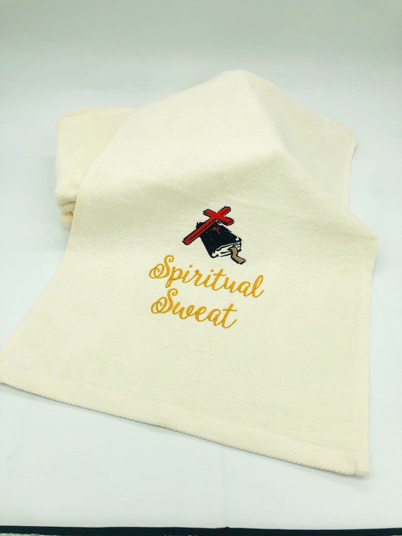 May include: A white hand towel with gold embroidery that reads "Spiritual Sweat". A small black and red illustration of a book with a cross on top is above the text.