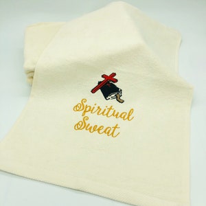 May include: A white towel with gold embroidery that reads "Spiritual Sweat". A red cross is embroidered above the text, and a black book is embroidered below the cross.
