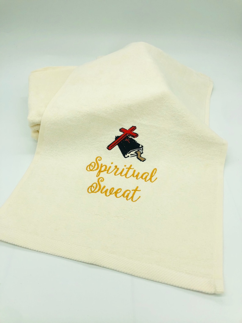 May include: A white towel with gold embroidery that reads "Spiritual Sweat". A red cross is embroidered above the text, and a black book is embroidered below the cross.