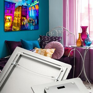 May include: A metal print of a colorful cityscape at night with a tall tower in the center. The print is framed with a silver frame and has a hanging wire on the back. The print is displayed on a white wall in a room with a bed and a window.