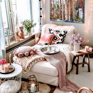 May include: A cozy living room with a white couch, a pink throw blanket, and a white knitted throw pillow. There is a painting of a birch forest on the wall, a small wooden stool, and a white side table with a candle on it. The room is decorated with fairy lights and flowers.