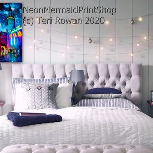 May include: A white bed with a gray headboard and a white duvet cover. The bed is decorated with a gray knitted throw blanket, a blue pillow, and a striped pillow. There is a white nightstand on the right side of the bed with a lamp and a picture frame. The wall behind the bed is white and has a string of lights hanging from it. A painting of a city street scene with a blue and purple sky is hanging on the wall above the nightstand. The painting is titled "NeonMermaidPrintShop (c) Teri Rowan 2020".