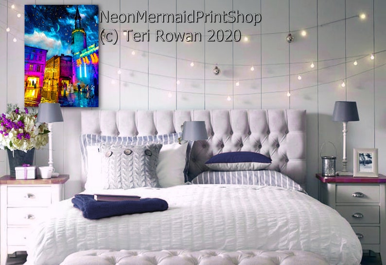 May include: A white bed with a gray headboard and a white duvet cover. The bed is decorated with a gray knitted throw blanket, a blue pillow, and a striped pillow. There is a white nightstand on the right side of the bed with a lamp and a picture frame. The wall behind the bed is white and has a string of lights hanging from it. A painting of a city street scene with a blue and purple sky is hanging on the wall above the nightstand. The painting is titled "NeonMermaidPrintShop (c) Teri Rowan 2020".