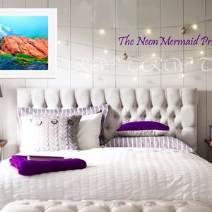 May include: A white bed with a tufted headboard and white bedding. The bed has a purple throw blanket and a grey knitted pillow. There is a framed picture of a mountain landscape above the headboard. The text "The Neon Mermaid Print Shop" is visible above the picture.