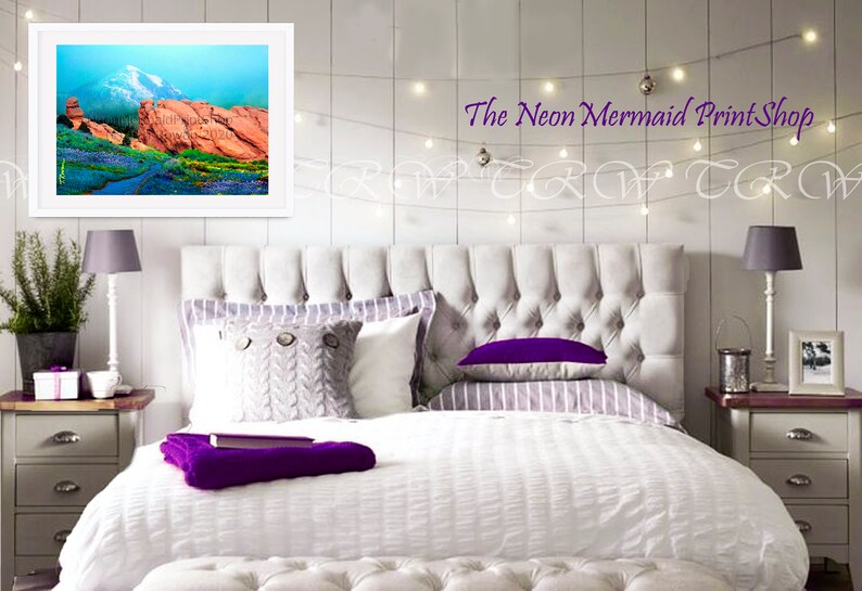 May include: A white bed with a tufted headboard and white bedding. The bed has a purple throw blanket and a grey knitted pillow. There is a framed picture of a mountain landscape above the headboard. The text "The Neon Mermaid Print Shop" is visible above the picture.