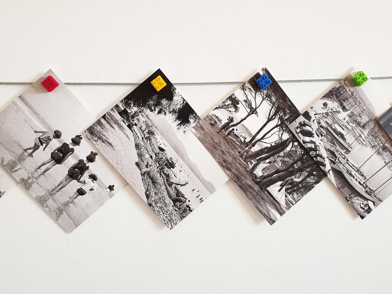 May include: Five black and white photographs are hanging on a white wall with colorful plastic clips. The photos are of different scenes, including a beach with people, a forest, and a harbor.