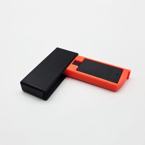 Könnte beinhalten: Eine schwarze rechteckige Hülle mit strukturierter Oberfläche ist geöffnet und zeigt einen orangefarbenen Halter mit einem schwarzen rechteckigen Objekt im Inneren. Der Halter hat an einem Ende eine kleine Kerbe. Die Artikel befinden sich auf weißem Hintergrund.