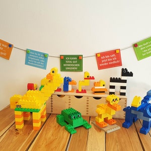 May include: A collection of colorful Lego animal figures, including a giraffe, a frog, a zebra, and two ducks. The figures are arranged on a wooden table with a wooden box behind them. There are also several colorful cards hanging from a string above the figures, each with a different message written in German.