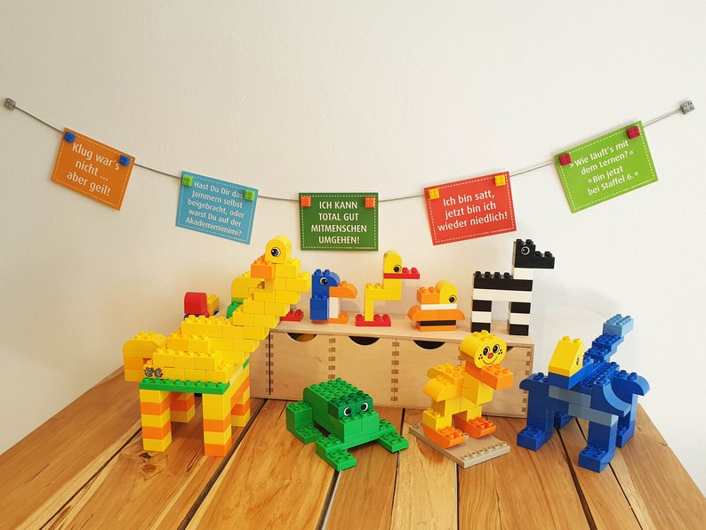 May include: A collection of colorful Lego animal figures, including a giraffe, a frog, a zebra, and two ducks. The figures are arranged on a wooden table with a wooden box behind them. There are also several colorful cards hanging from a string above the figures, each with a different message written in German.