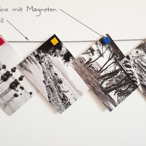 May include: Black and white photos are hung on a white wall with a thin wire. The photos are held in place with colorful plastic building blocks. The text "Bausteine mit Magneten" and "Stahlseil" is written on the wall.