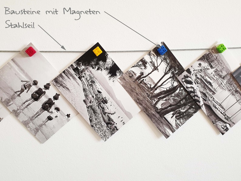 May include: Black and white photos are hung on a white wall with a thin wire. The photos are held in place with colorful plastic building blocks. The text "Bausteine mit Magneten" and "Stahlseil" is written on the wall.