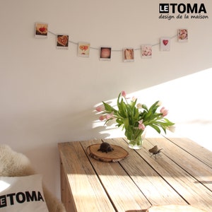 Op de afbeelding: Een houten tafel met een vaas met roze tulpen, een klein houten schijfje met een decoratieve kever en een kleine metalen vlinder. De tafel staat voor een witte muur met een string met foto's die eraan hangen. De foto's zijn van harten, snoep en andere romantische thema's. De tekst "LE TOMA design de la maison" is op de muur gedrukt.